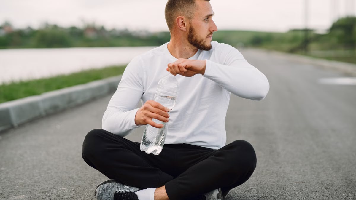 Man seated on an empty road holding a water bottle, focusing on healthy lifestyle.
