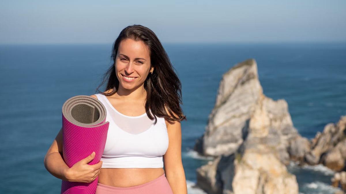 Smiling woman with yoga mat in sportswear by oceanic cliffs in Portugal.